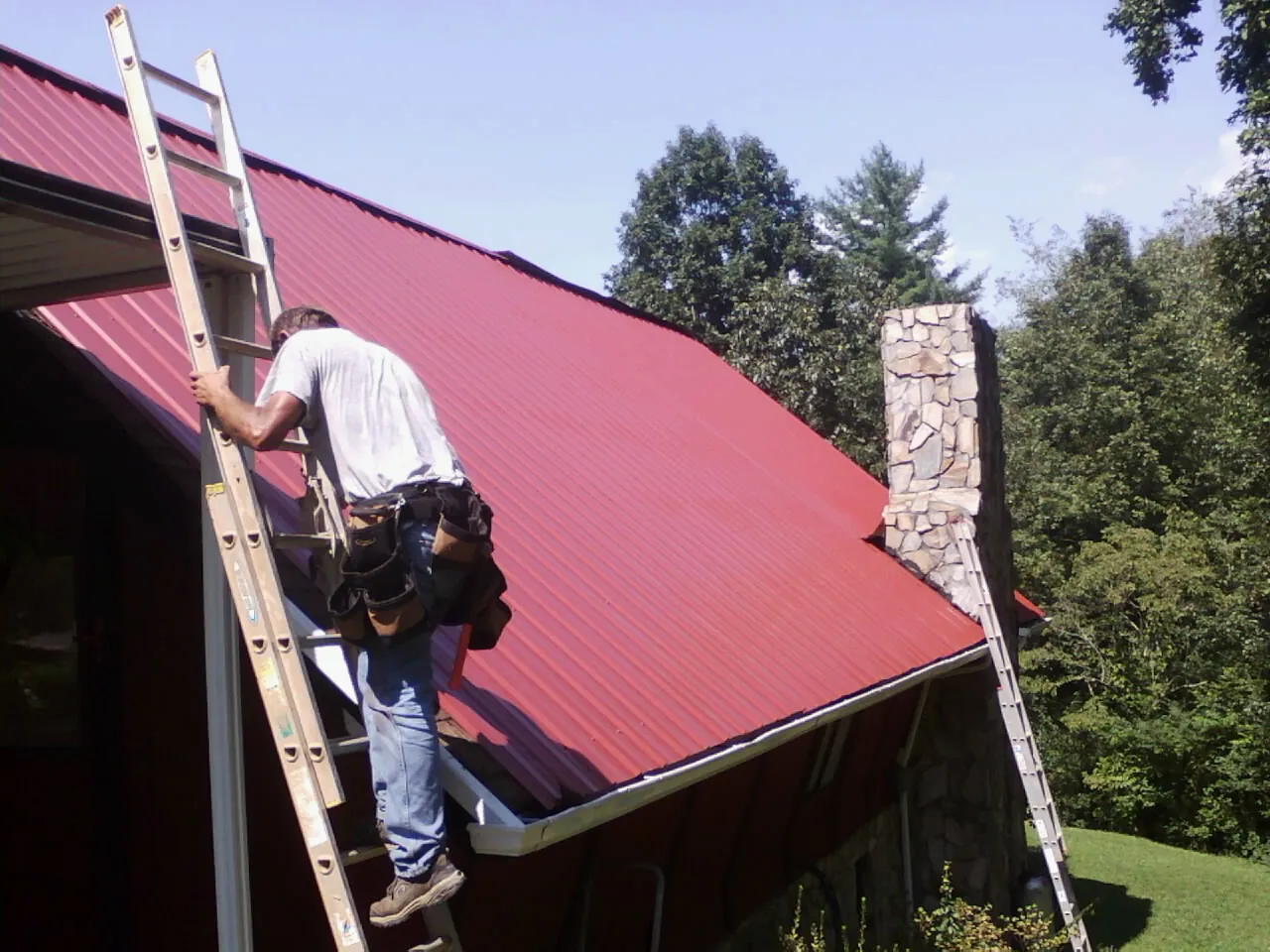 Roofer on a ladder beside a red metal roof and stone chimney — Soffit & Fascia Repair by Roofing Brothers in Cornwall On Hudson